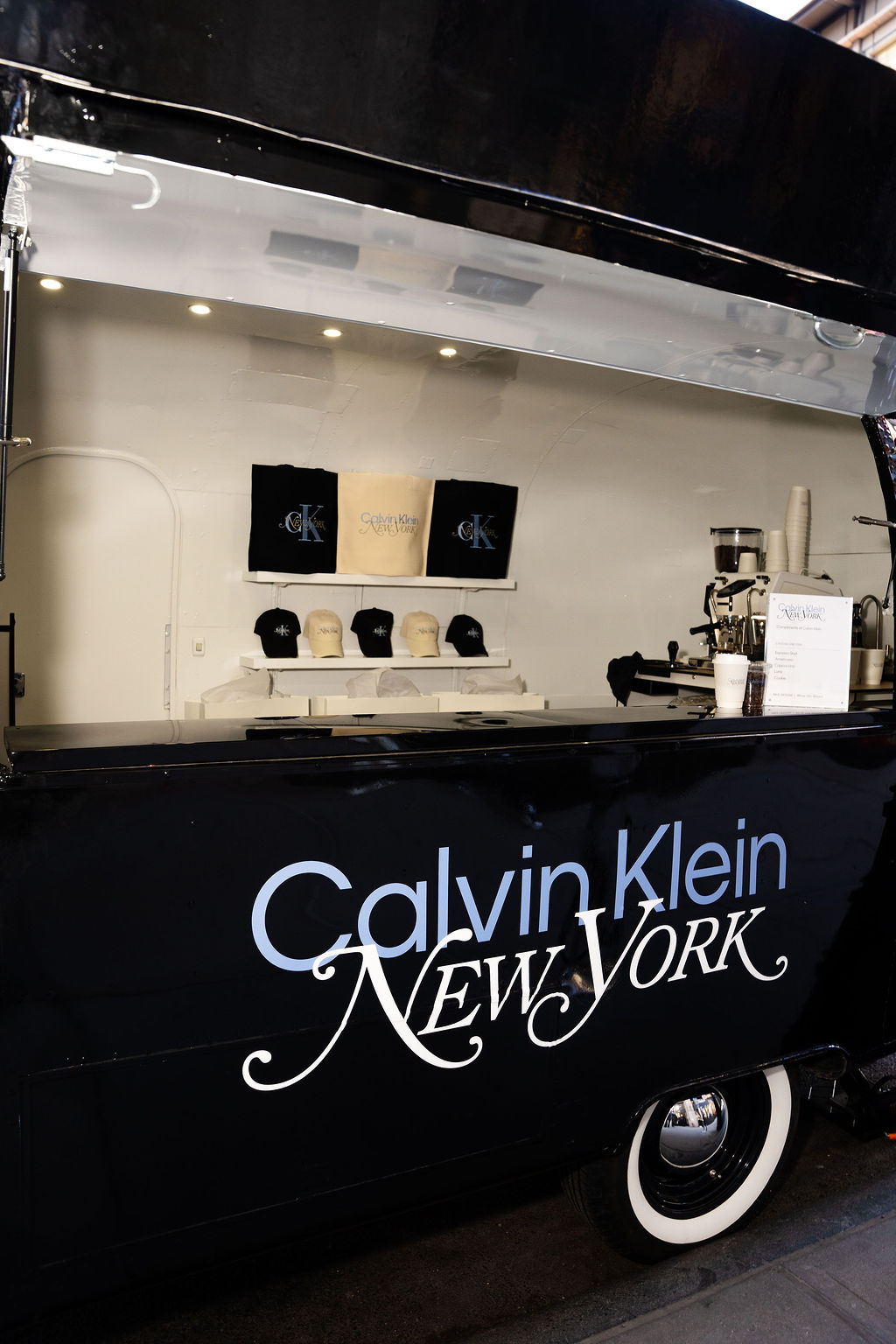 Black Calvin Klein food truck displays branded hats, mugs, shirts, coffee machine, and cups.