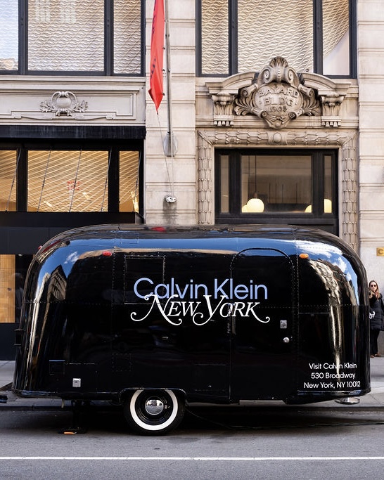 A black trailer labeled Calvin Klein New York is parked on a city street by a large stone building.