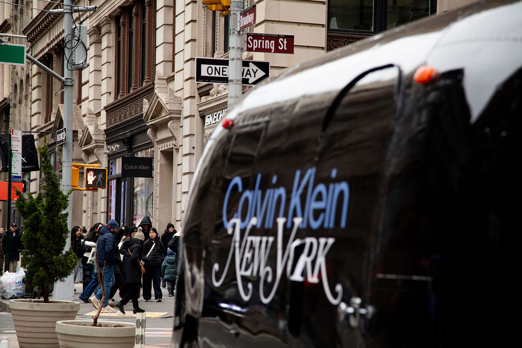 People cross a New York street with a Calvin Klein van and signs for Spring St and One Way visible.