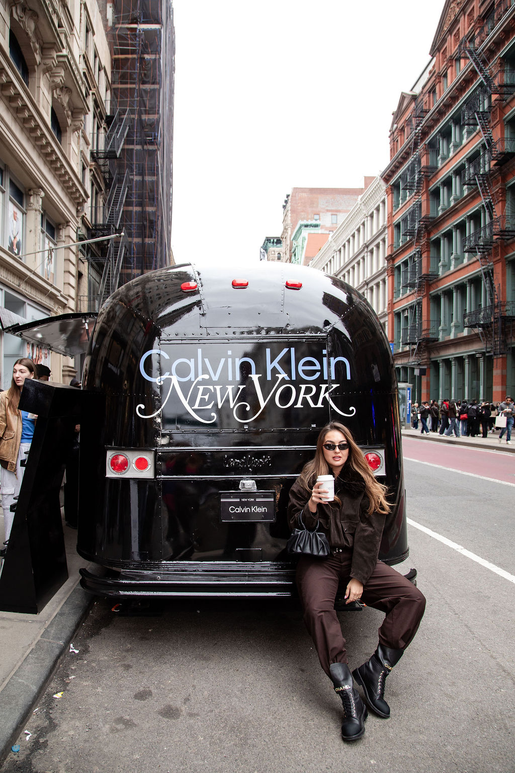 Woman in sunglasses sits on a black Calvin Klein trailer, holding coffee on a city street.