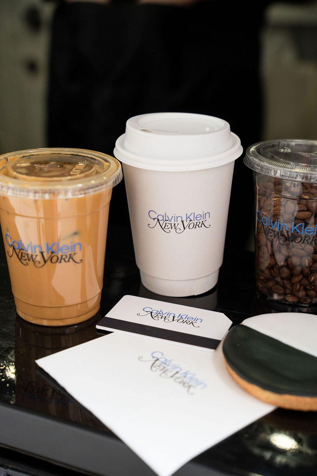 Three Calvin Klein cups with iced coffee, hot coffee, and beans, plus cards, cookie, and napkin.
