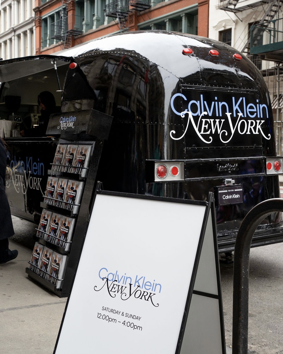 Black Calvin Klein New York trailer on city street with magazines and sign showing weekend hours.