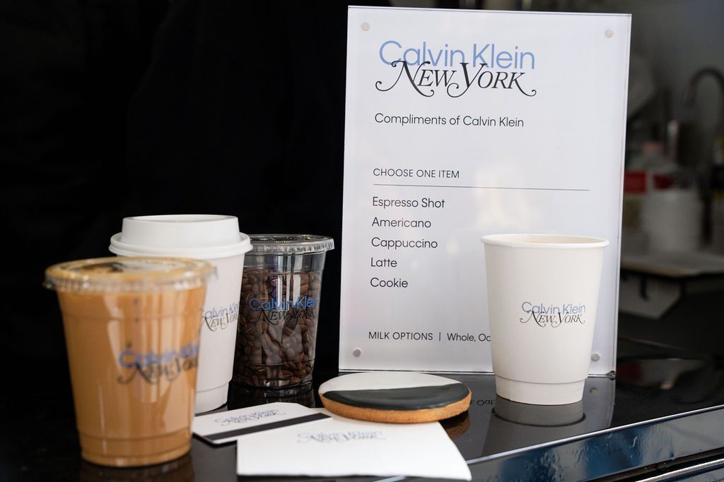 Calvin Klein cups with coffee, coffee beans, a cookie, napkins, and a free coffee sign.