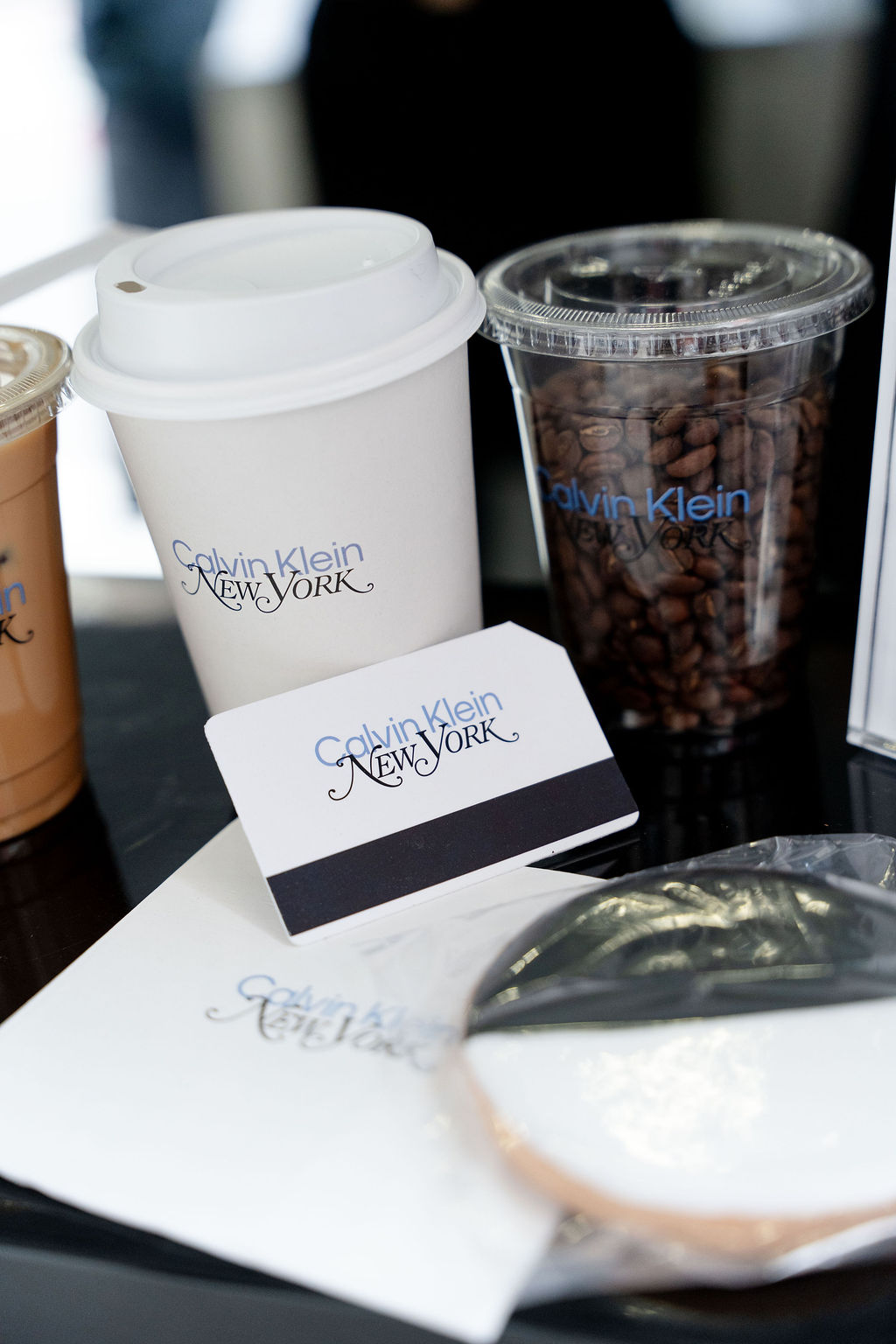 Calvin Klein New York coffee cup, cup of beans, and business card on table with drinks and napkin.