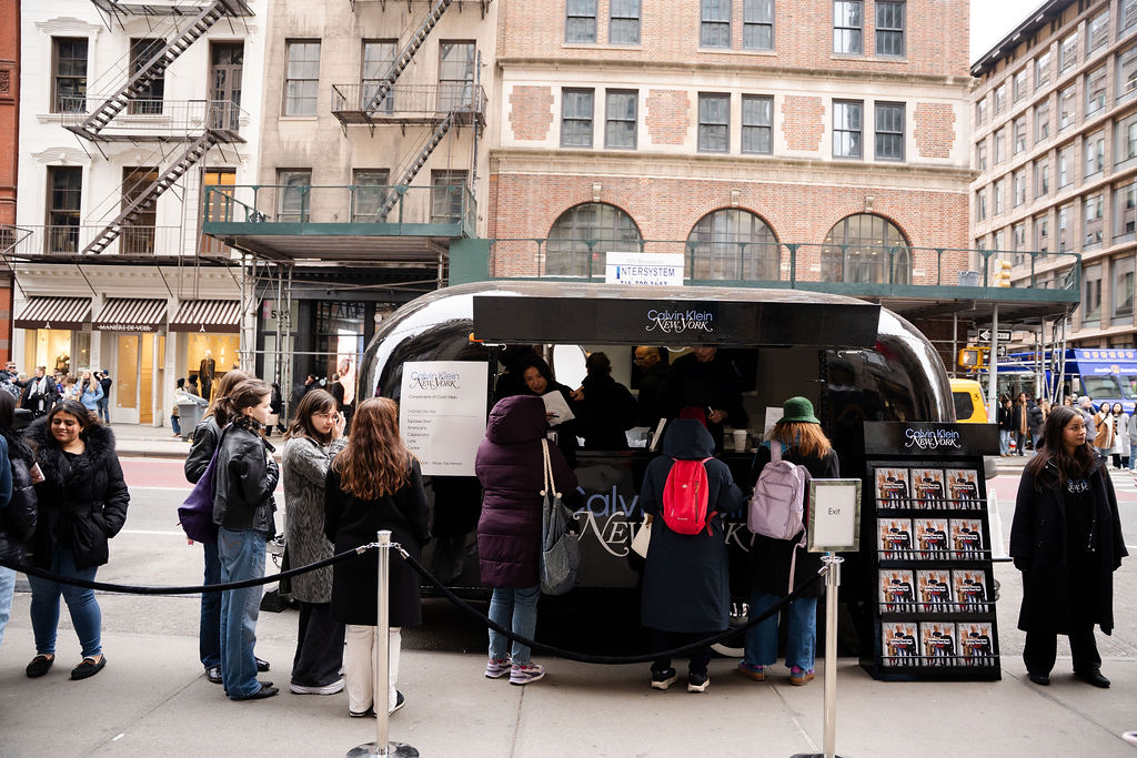 People line up at a black pop-up booth on a city street near tall buildings and a display stand.