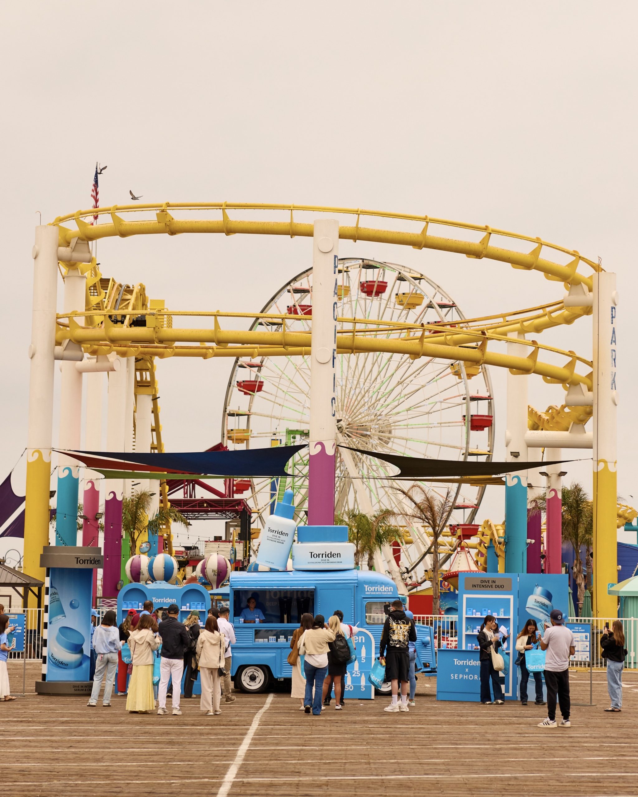 People line up at a blue food truck on a boardwalk with a roller coaster and Ferris wheel behind.