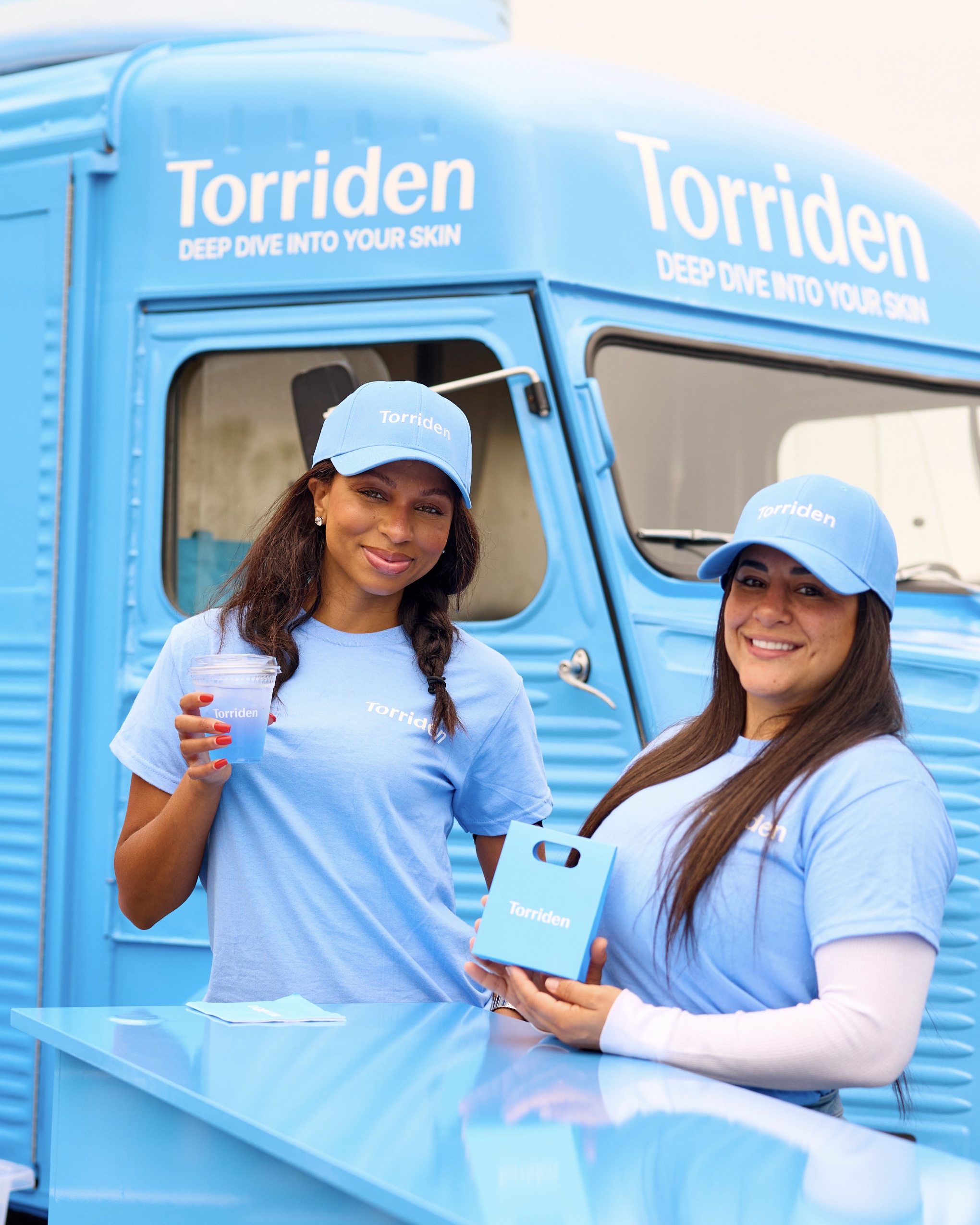 Two women in light blue Torriden hats and shirts stand by a blue Torriden truck, smiling.