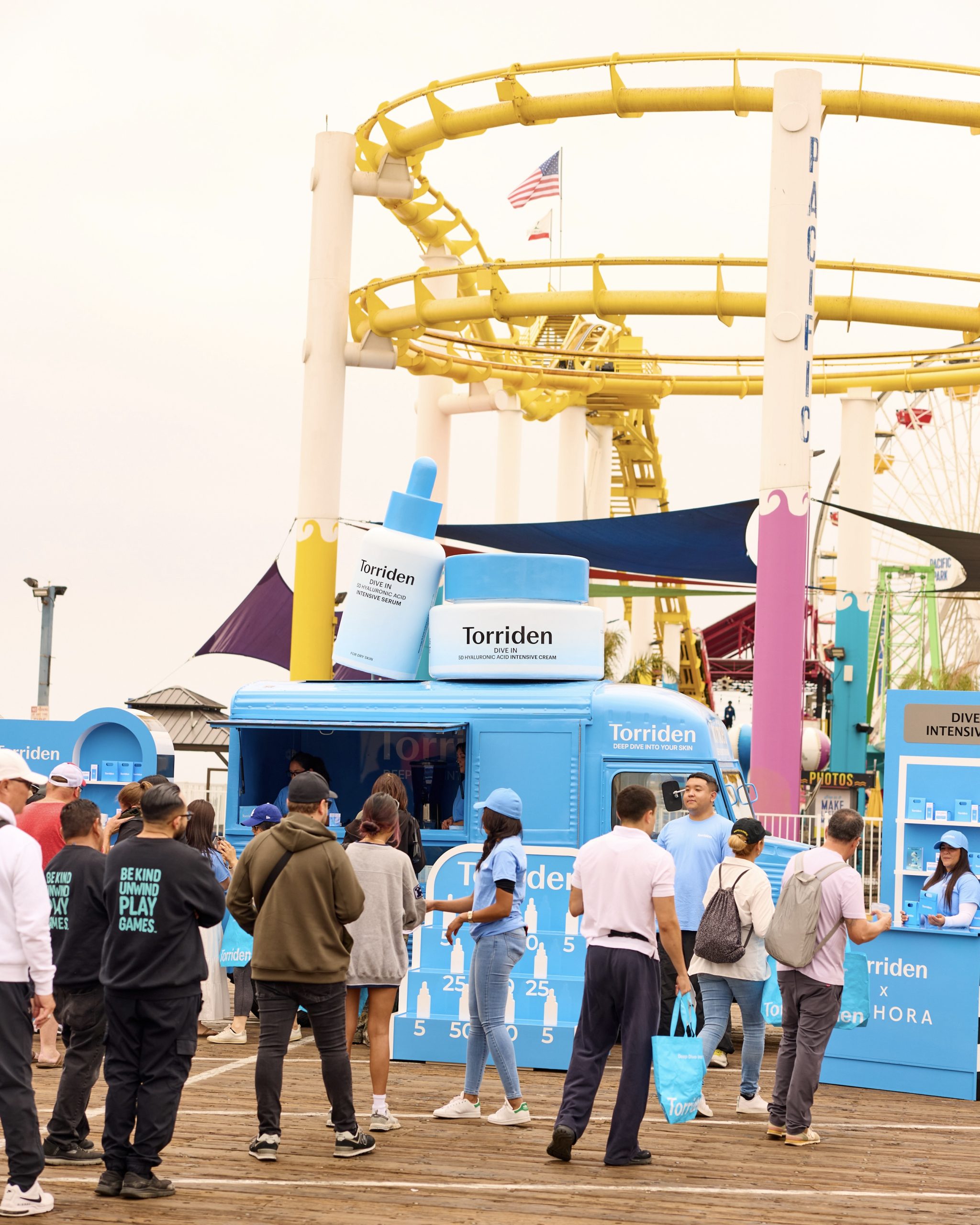 People crowd a blue Torriden skincare booth on a pier, with rides in the background.