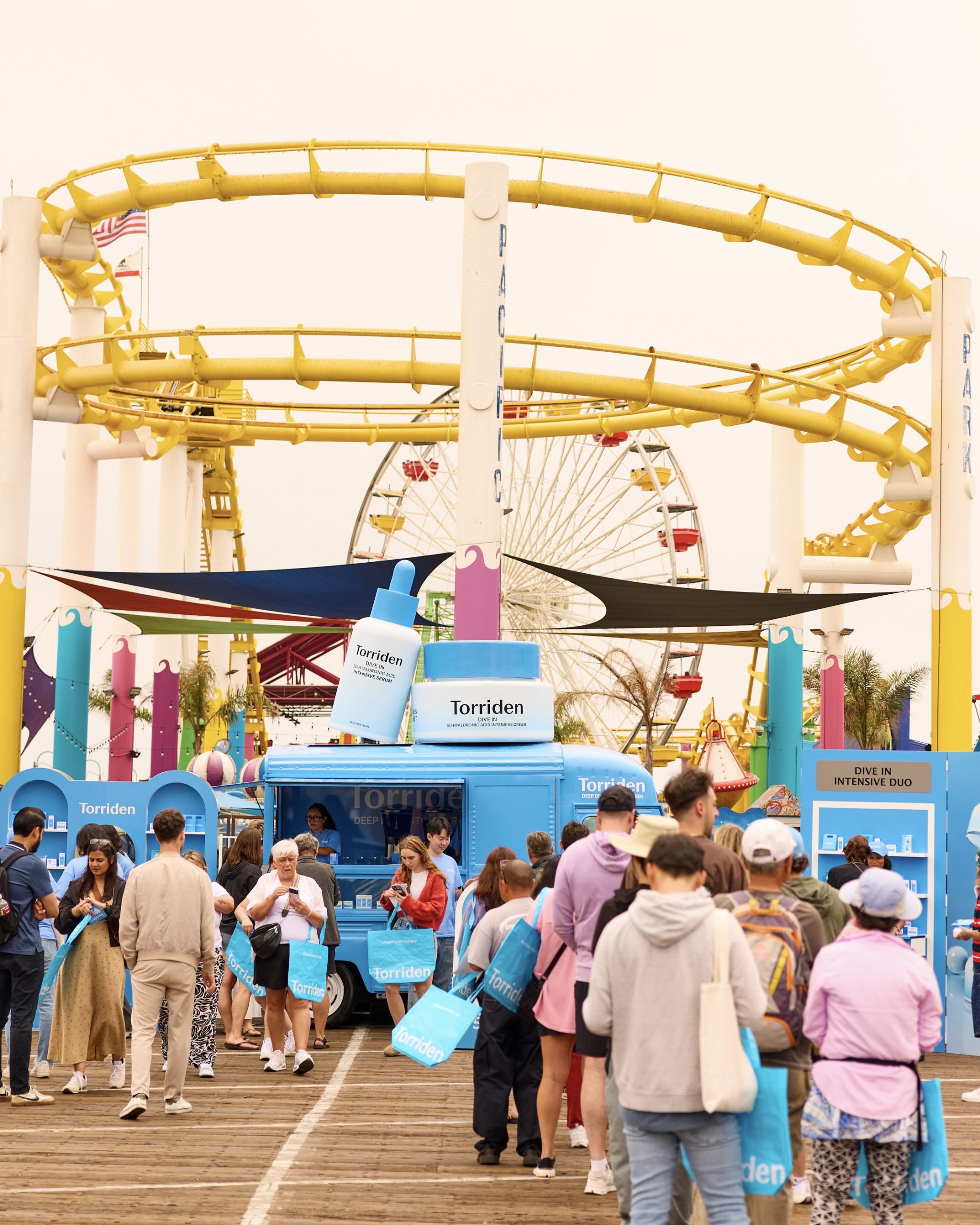 People gather at a blue Torriden pop-up truck on a pier with roller coaster and Ferris wheel nearby.