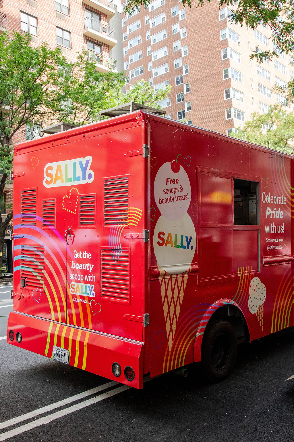 Red food truck labeled SALLY with ice cream and Pride designs, parked by tall city buildings.