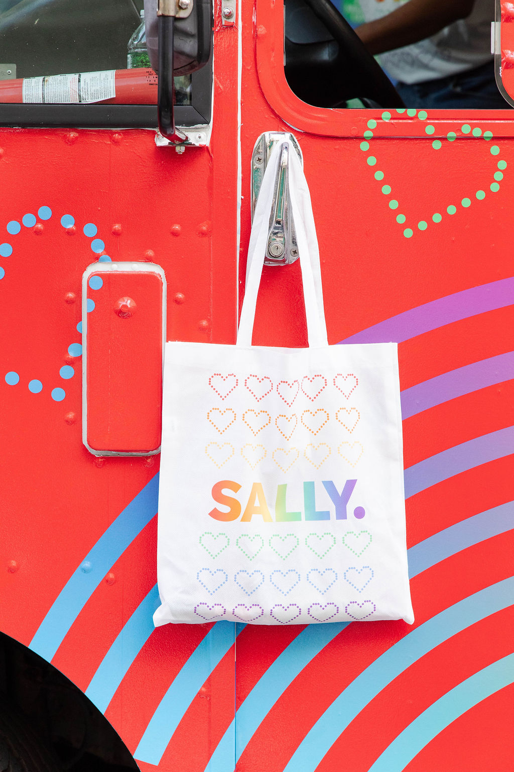 White tote bag with dotted hearts and rainbow "SALLY" hangs on red vehicle with pastel hearts.