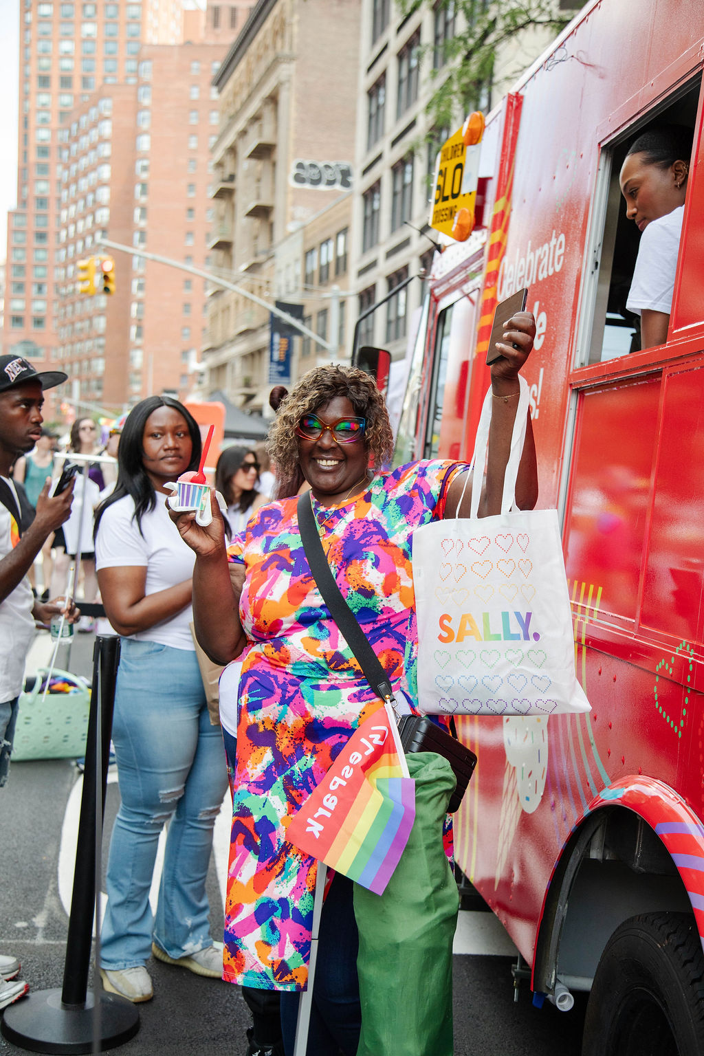 Person in a colorful dress holds ice cream and a tote bag by a red food truck at a city event.
