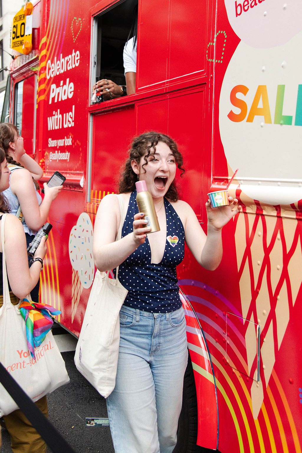 Young person with curly hair holds beauty products and a cup in front of a Pride truck with others nearby.
