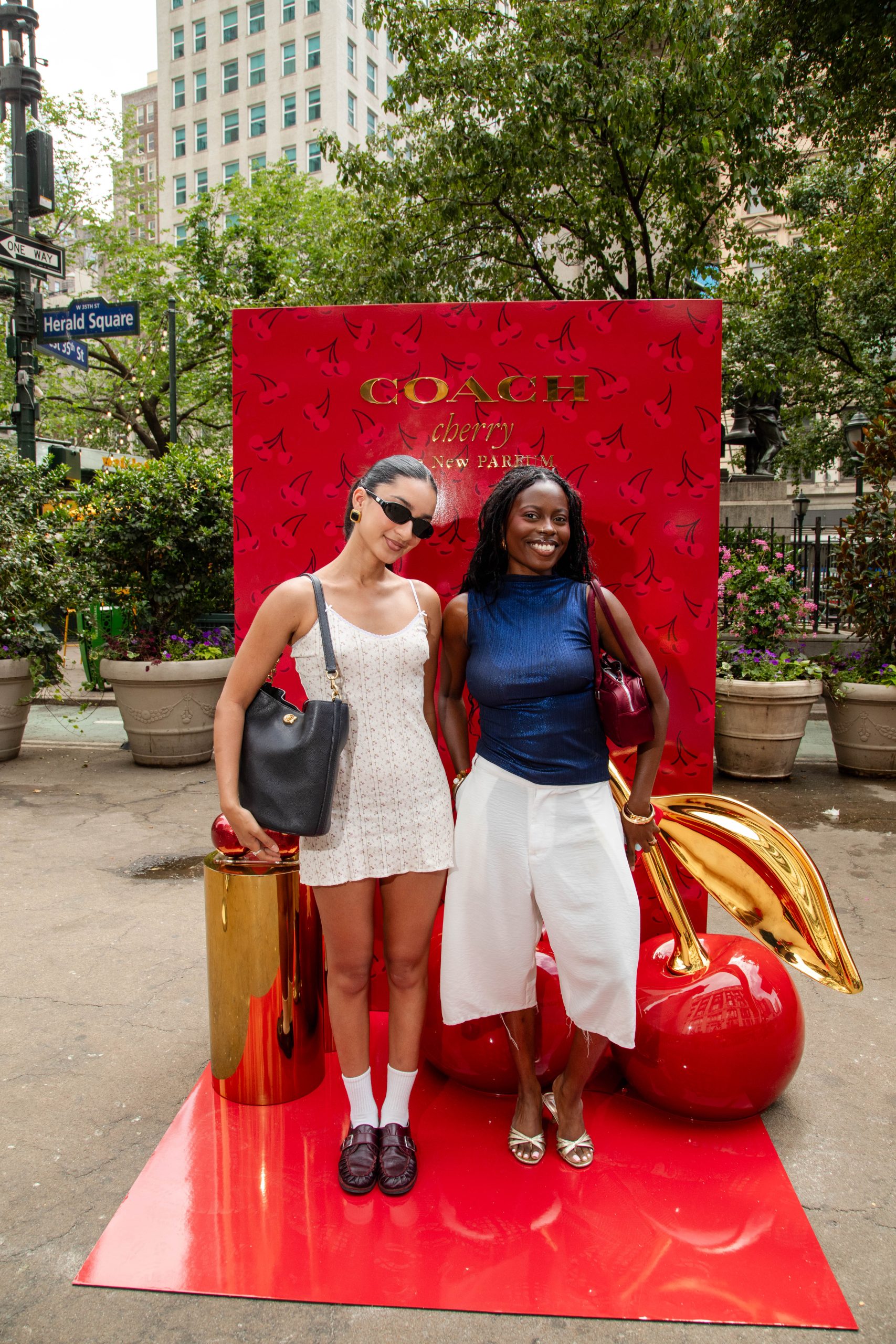 Two women smile in front of a red Coach Cherry perfume display with large cherries outdoors.