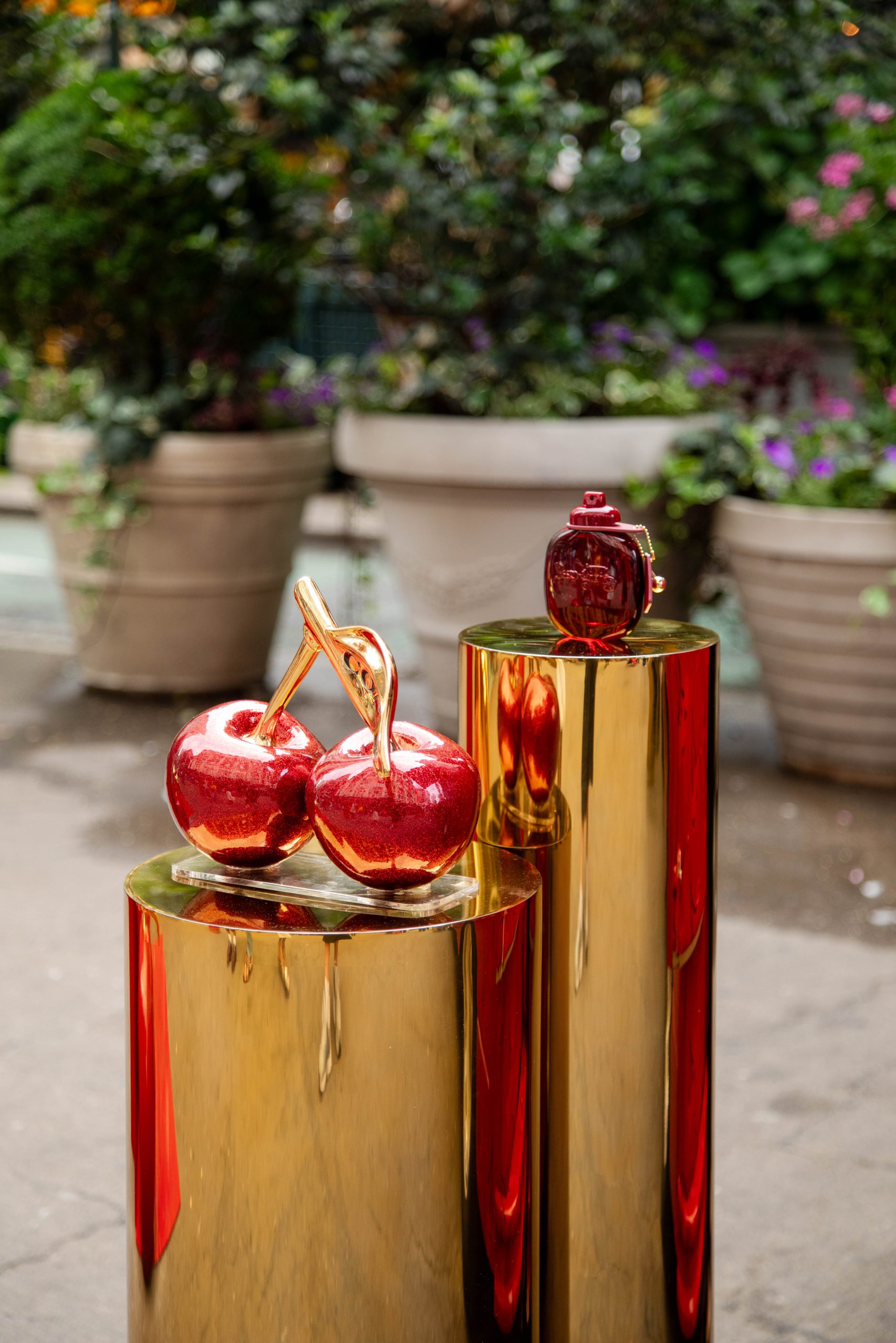 Red apple and grenade sculptures stand on gold pedestals outdoors with potted plants behind them.