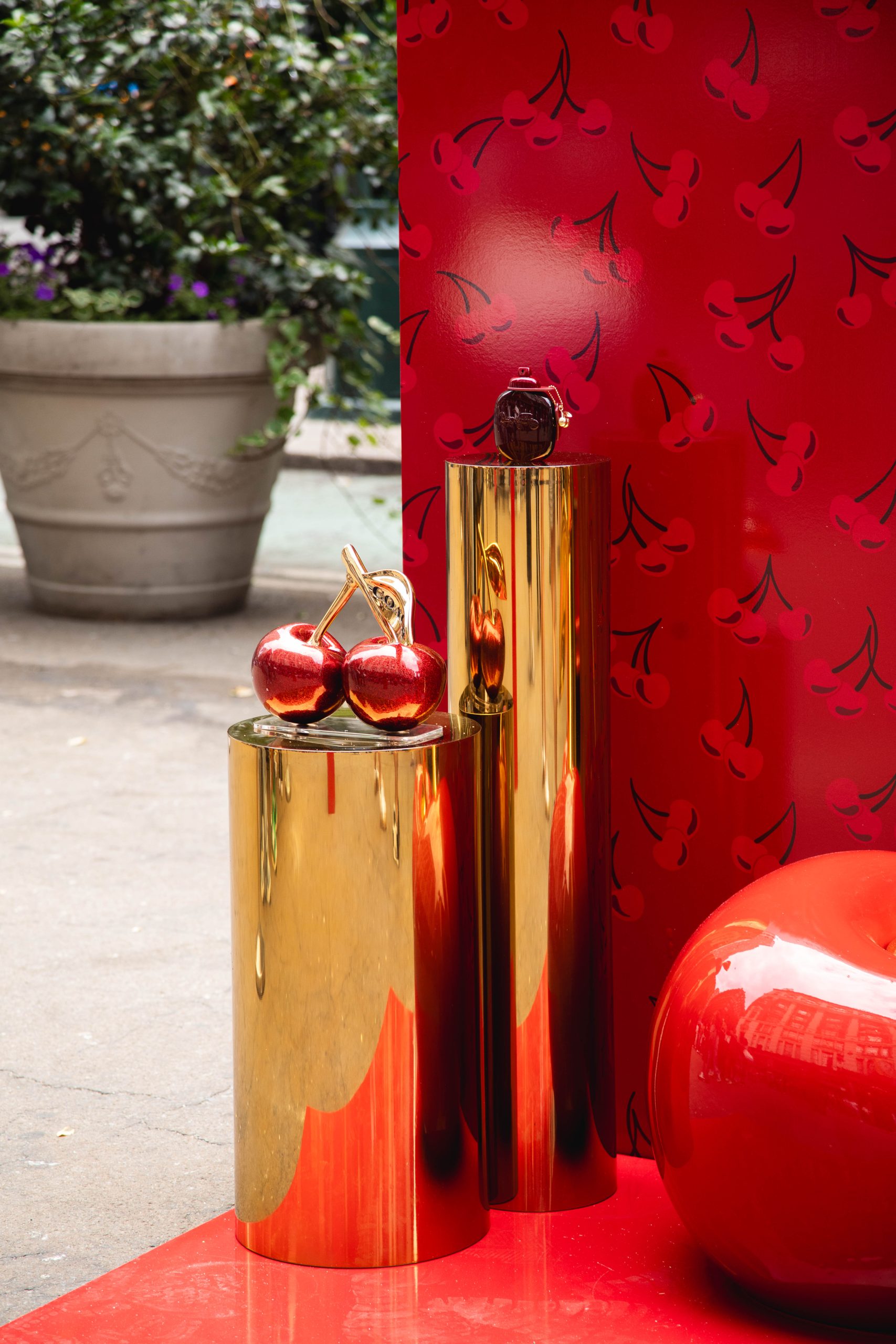 Gold pedestals with metallic cherry sculptures beside a red object and cherry-patterned backdrop.