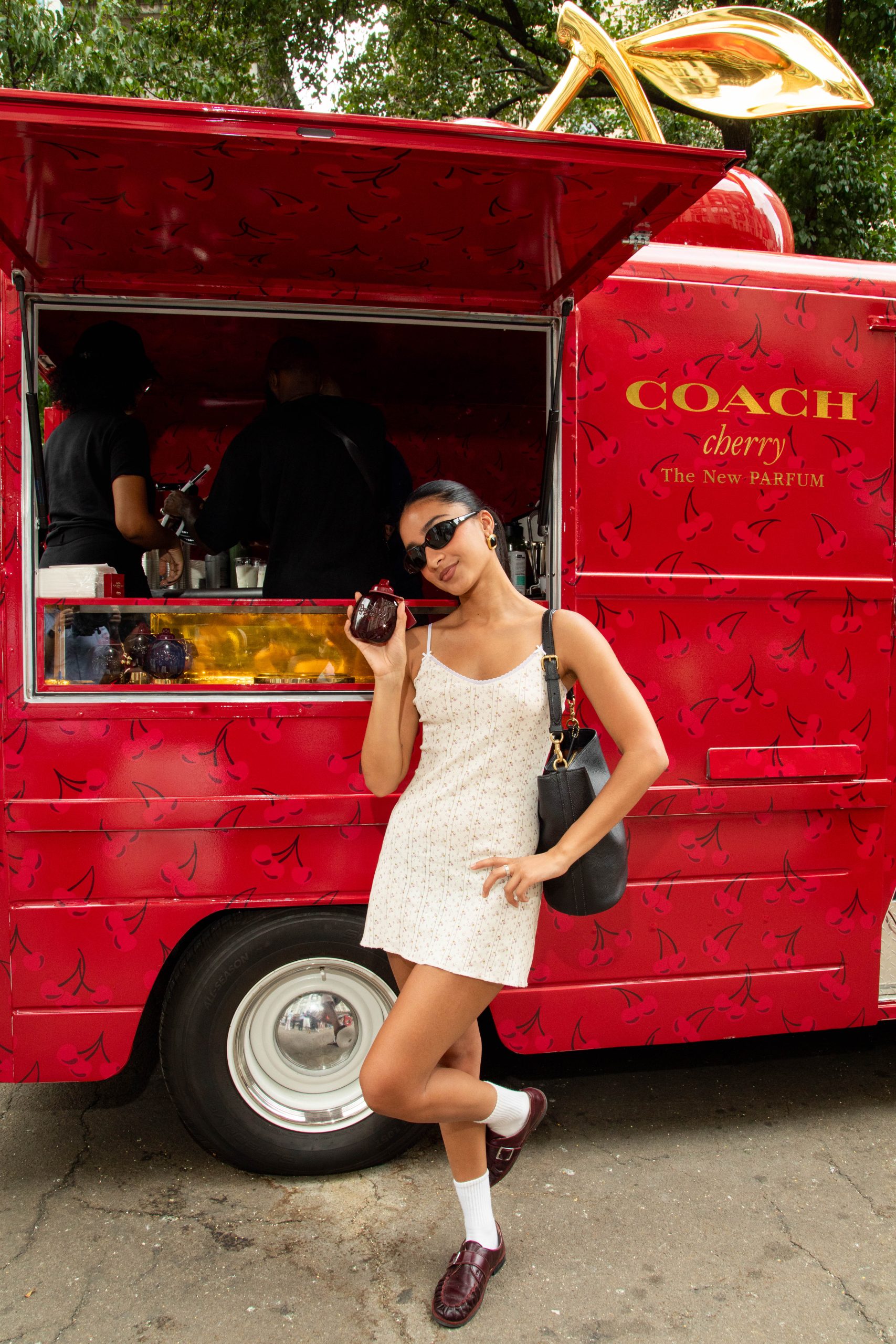 Woman in white dress holds perfume by a red Coach Cherry truck, wearing sunglasses and black bag.