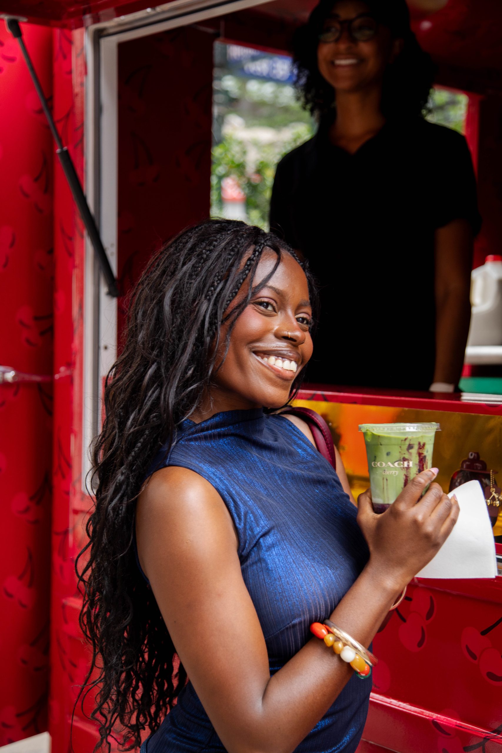 Woman in blue dress holds green drink and napkin by red food truck; another person smiles inside.