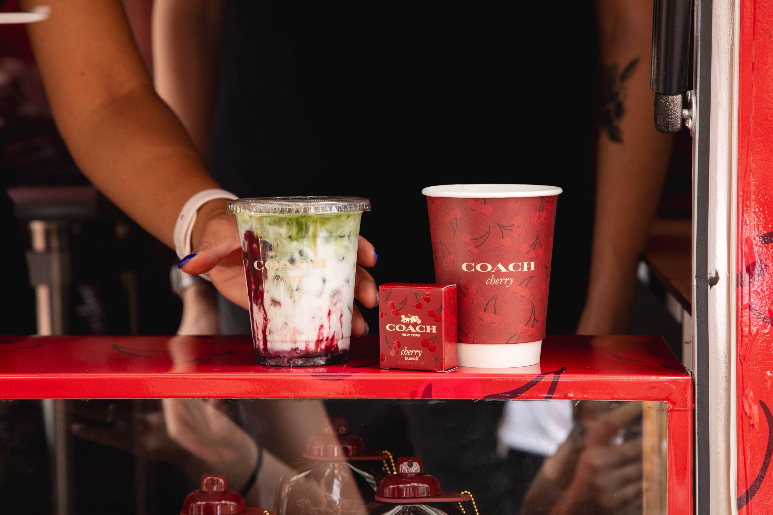 A person sets a matcha berry drink, red paper cup, and red Coach box on a red counter.