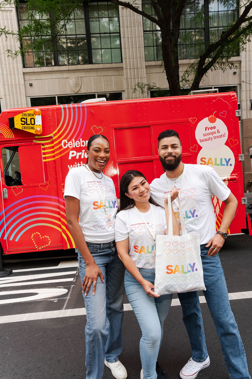 Three people in matching SALLY Pride shirts stand by a rainbow-decorated red truck with a tote bag.