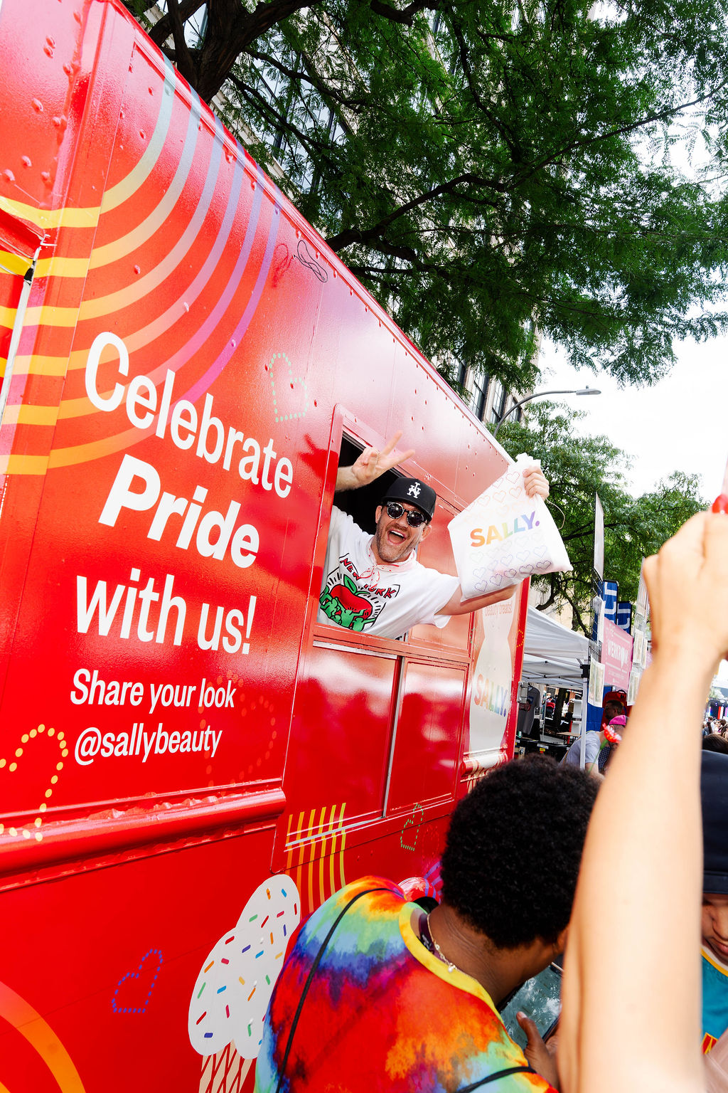 A person in sunglasses leans out of a red pride truck holding a bag as people cheer outside.