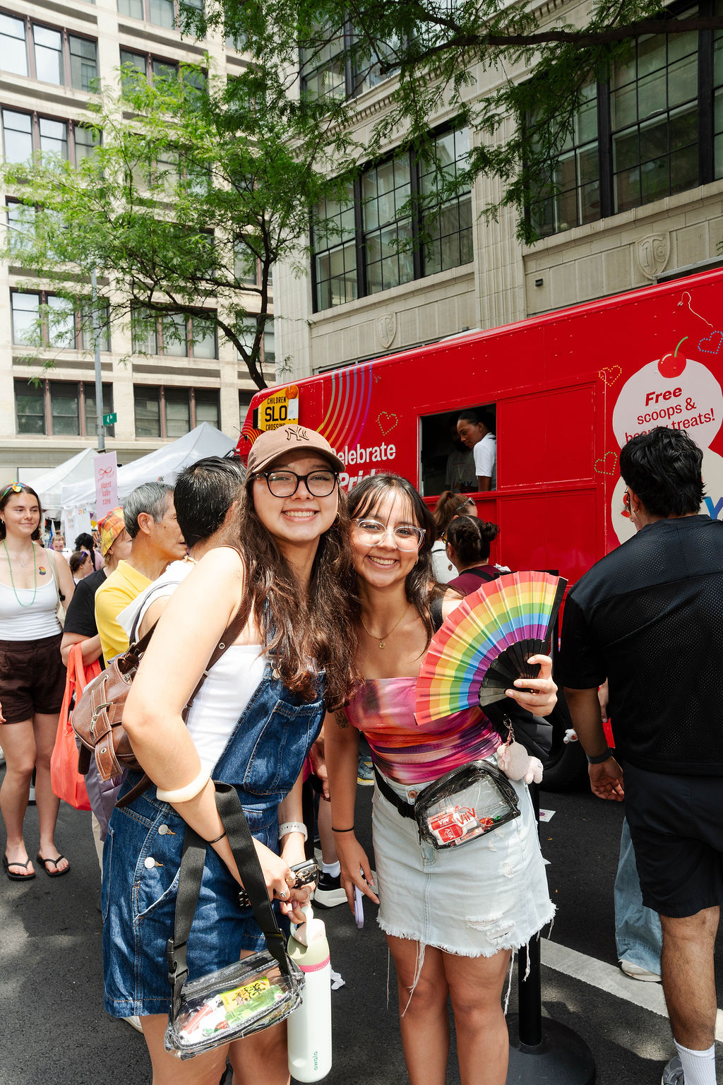 Two people smile at an outdoor event; one in overalls, the other with a rainbow fan. Red food truck behind.