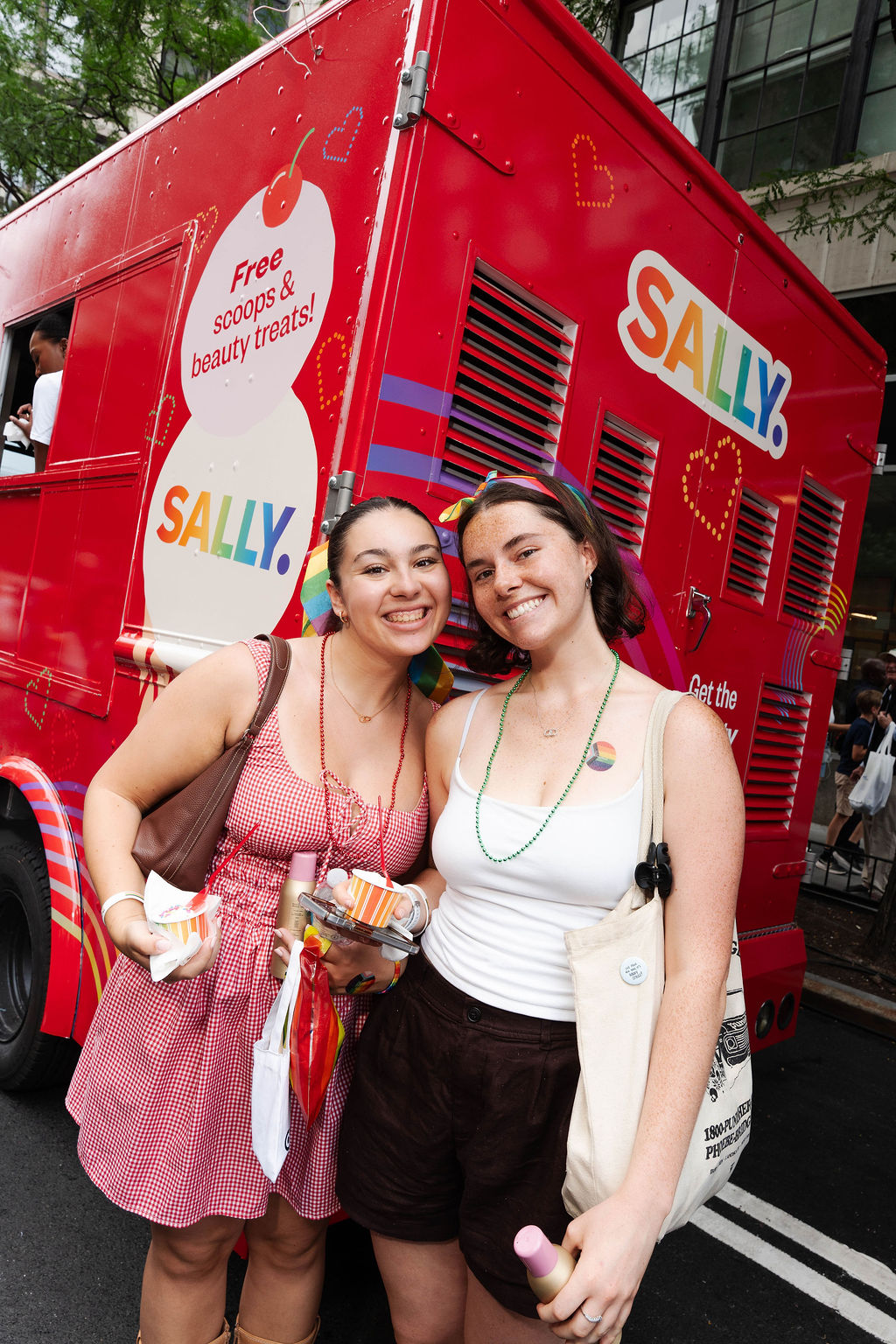 Two women with snacks and tote bags stand by a red SALLY. ice cream truck at an outdoor event.