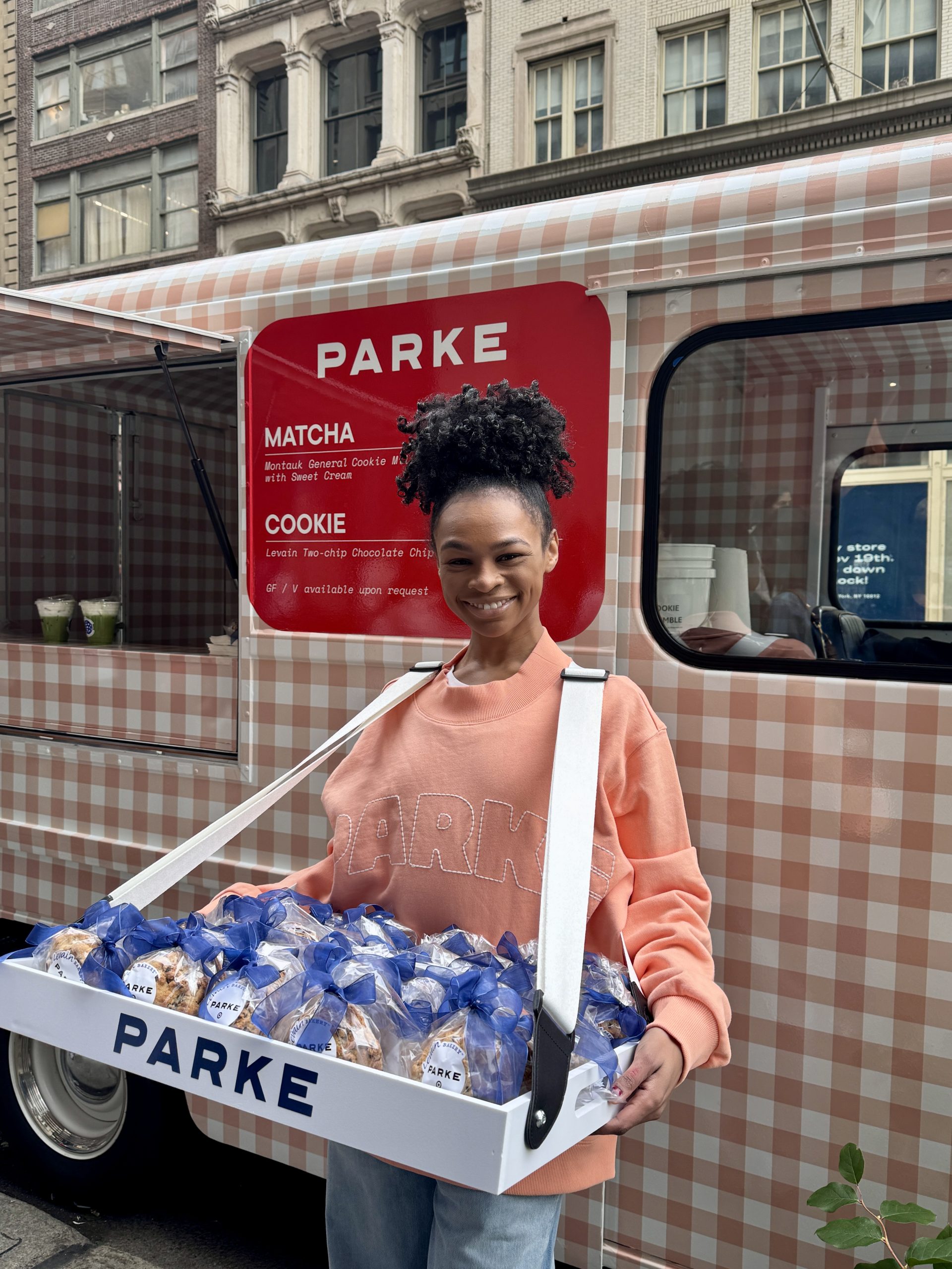 Woman in peach sweater holds a tray of blue-ribbon wrapped cookies by a checkered food truck.