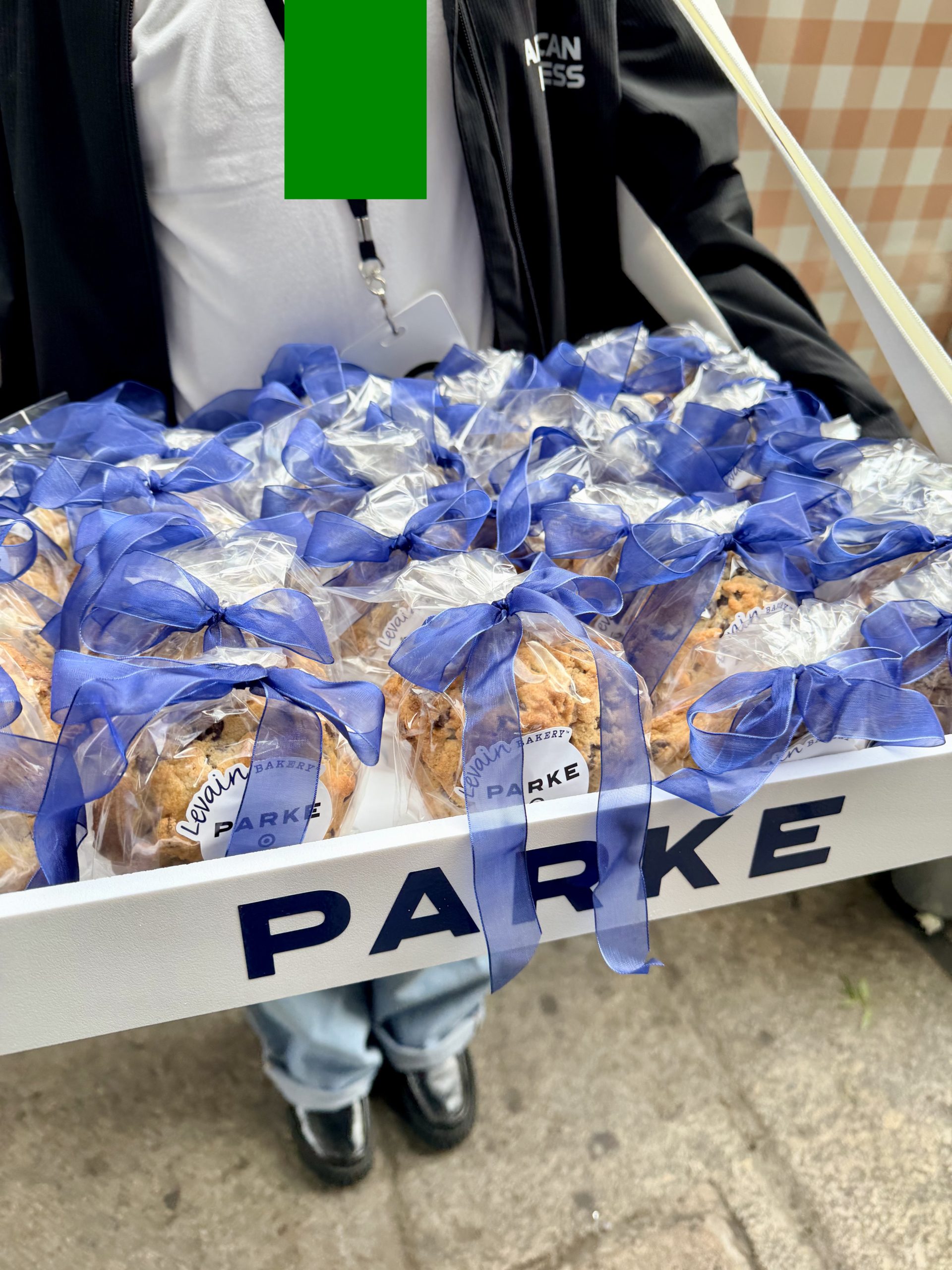 Person holds a white tray labeled PARKE with bagged cookies tied with blue ribbons outdoors.
