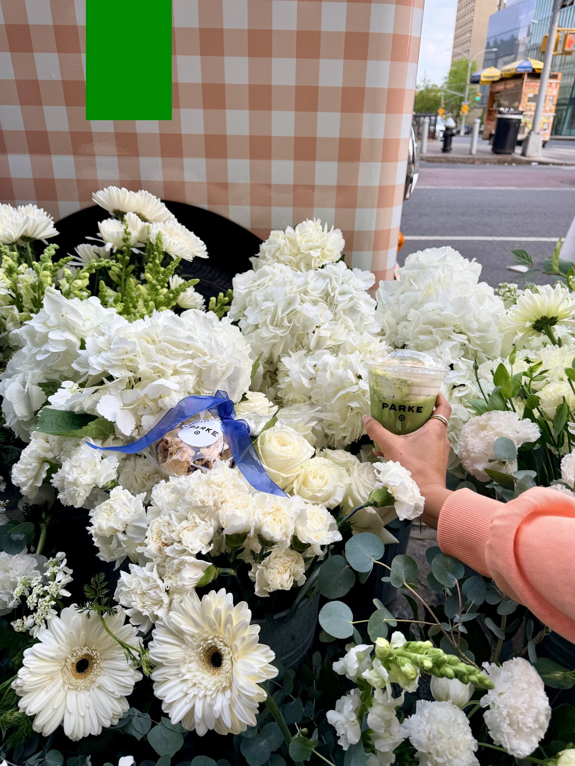Person in a peach sleeve holds a “PARKE” cup among white flowers at an outdoor market.