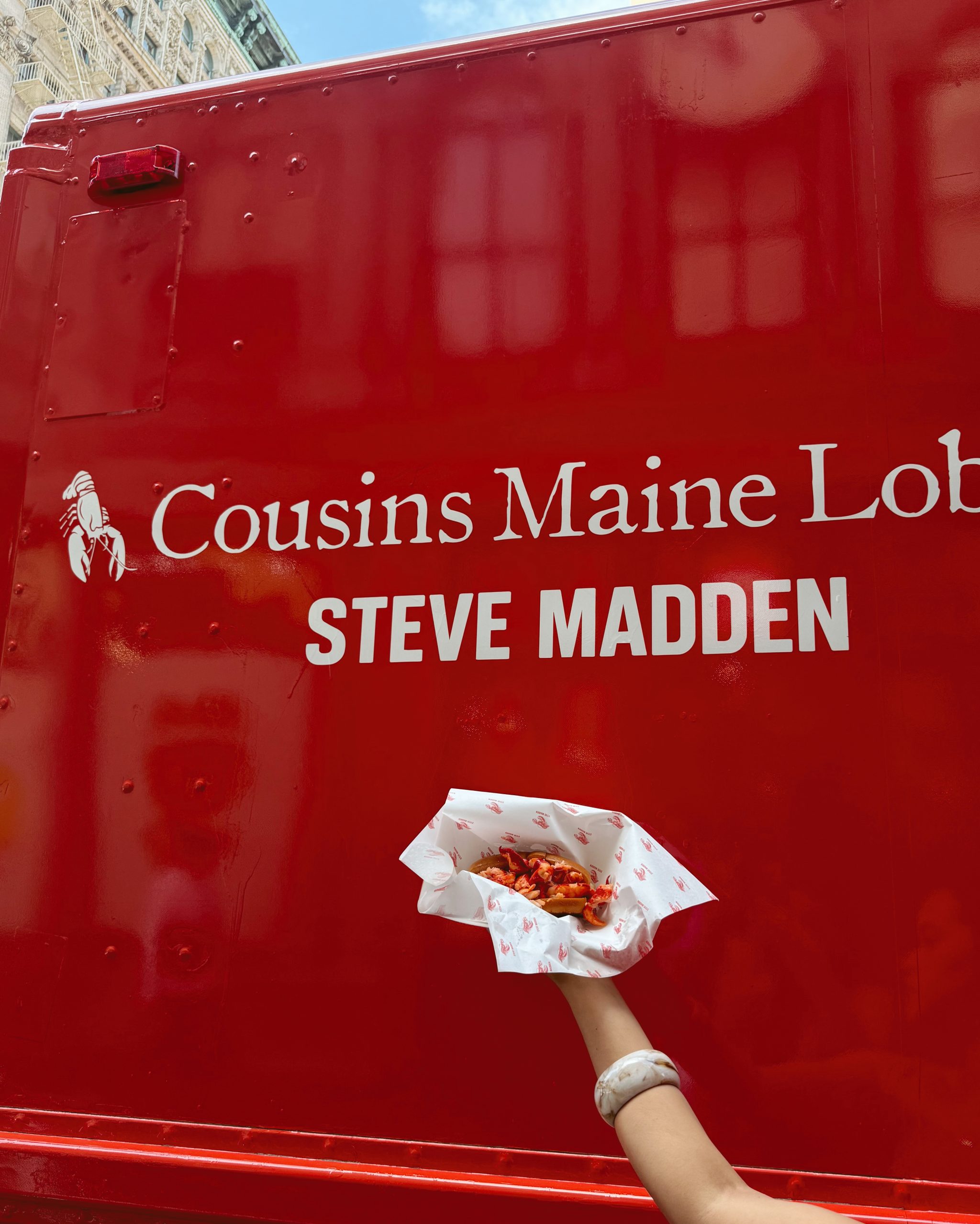 Hand holding lobster roll in front of red Cousins Maine Lobster truck with STEVE MADDEN sign.