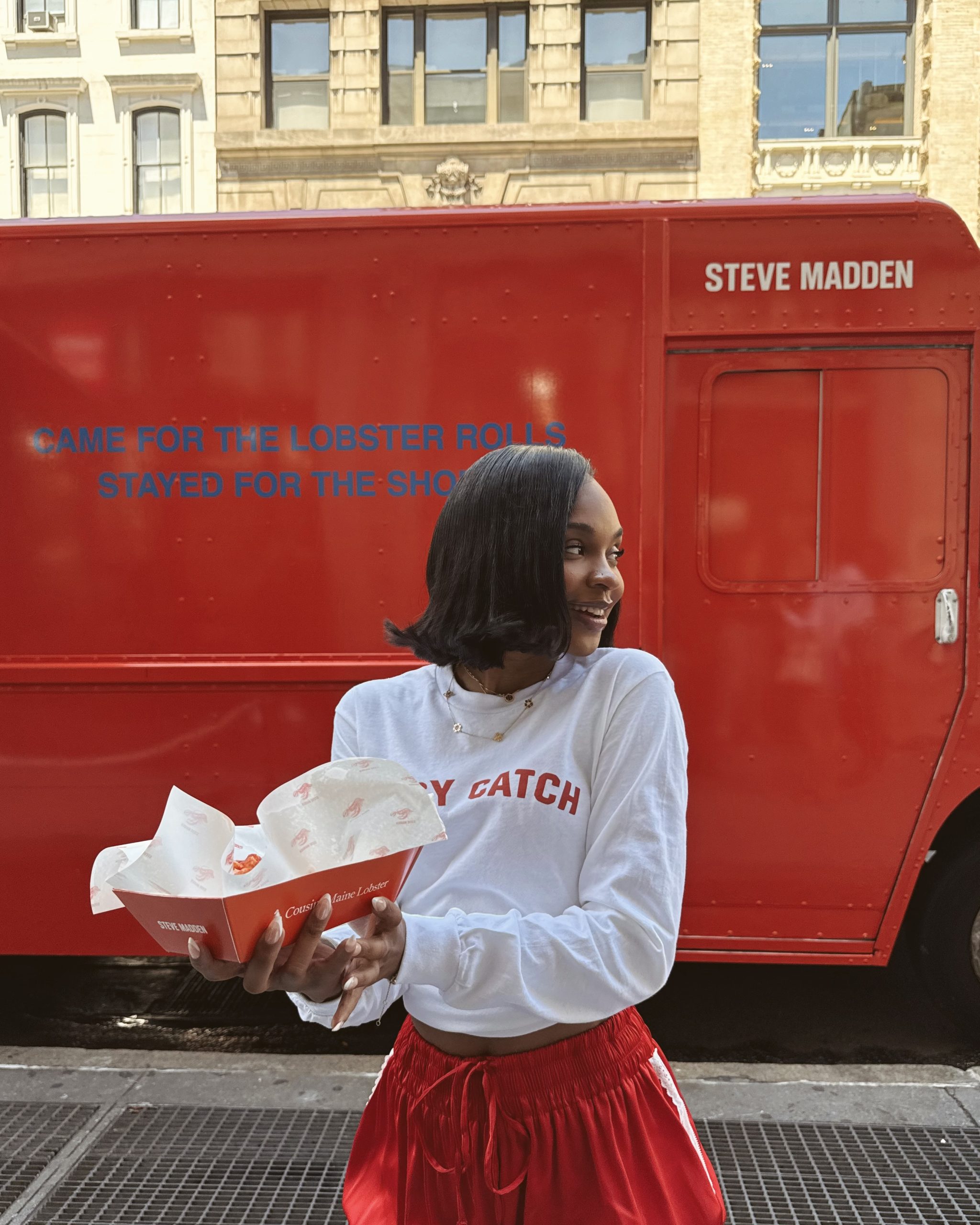 Smiling woman holding empty food containers in front of red STEVE MADDEN food truck with slogans.
