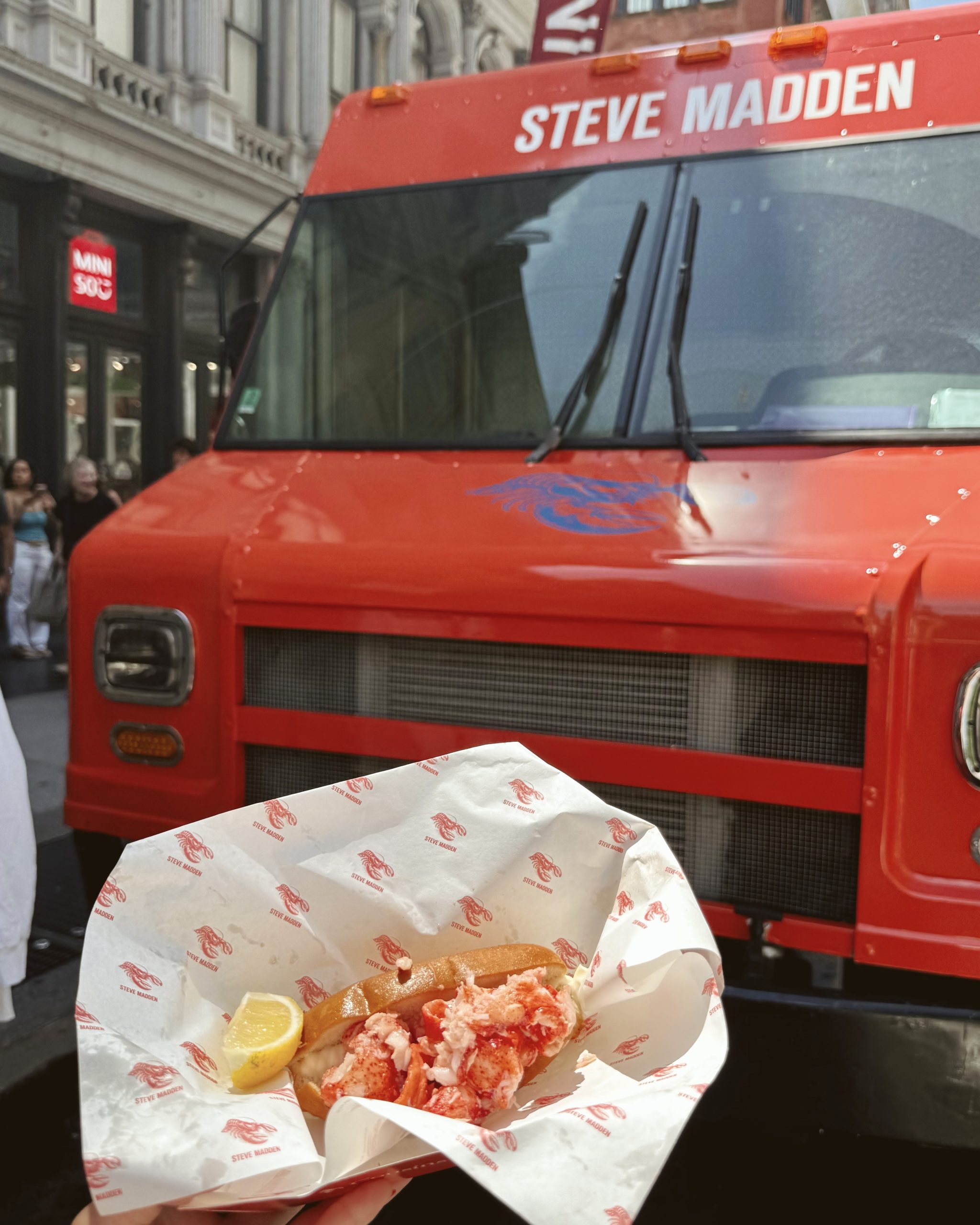 Lobster roll with lemon wedge held before a bright orange Steve Madden food truck.