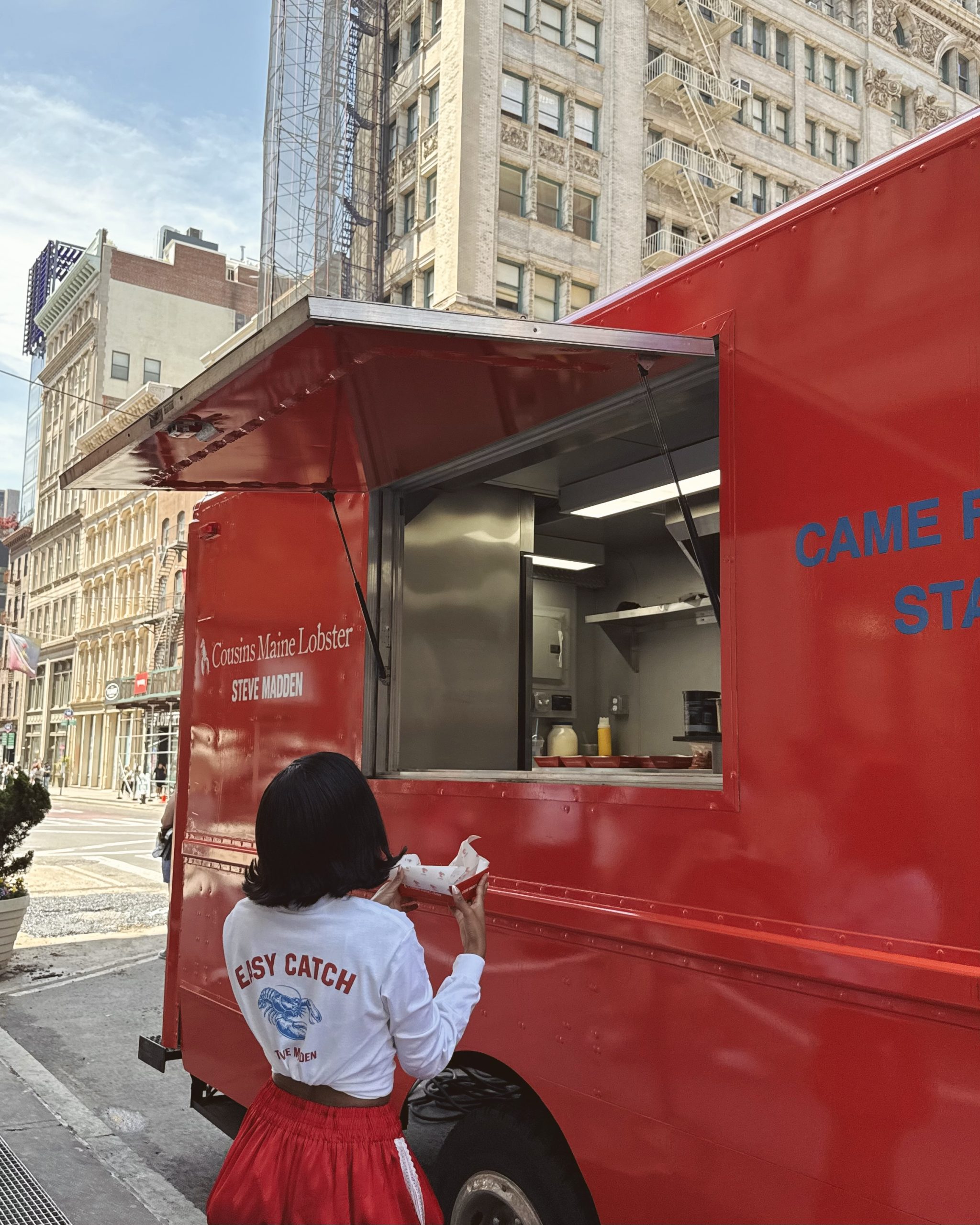 Person in white shirt and red skirt stands by a red food truck on a city street, holding food.