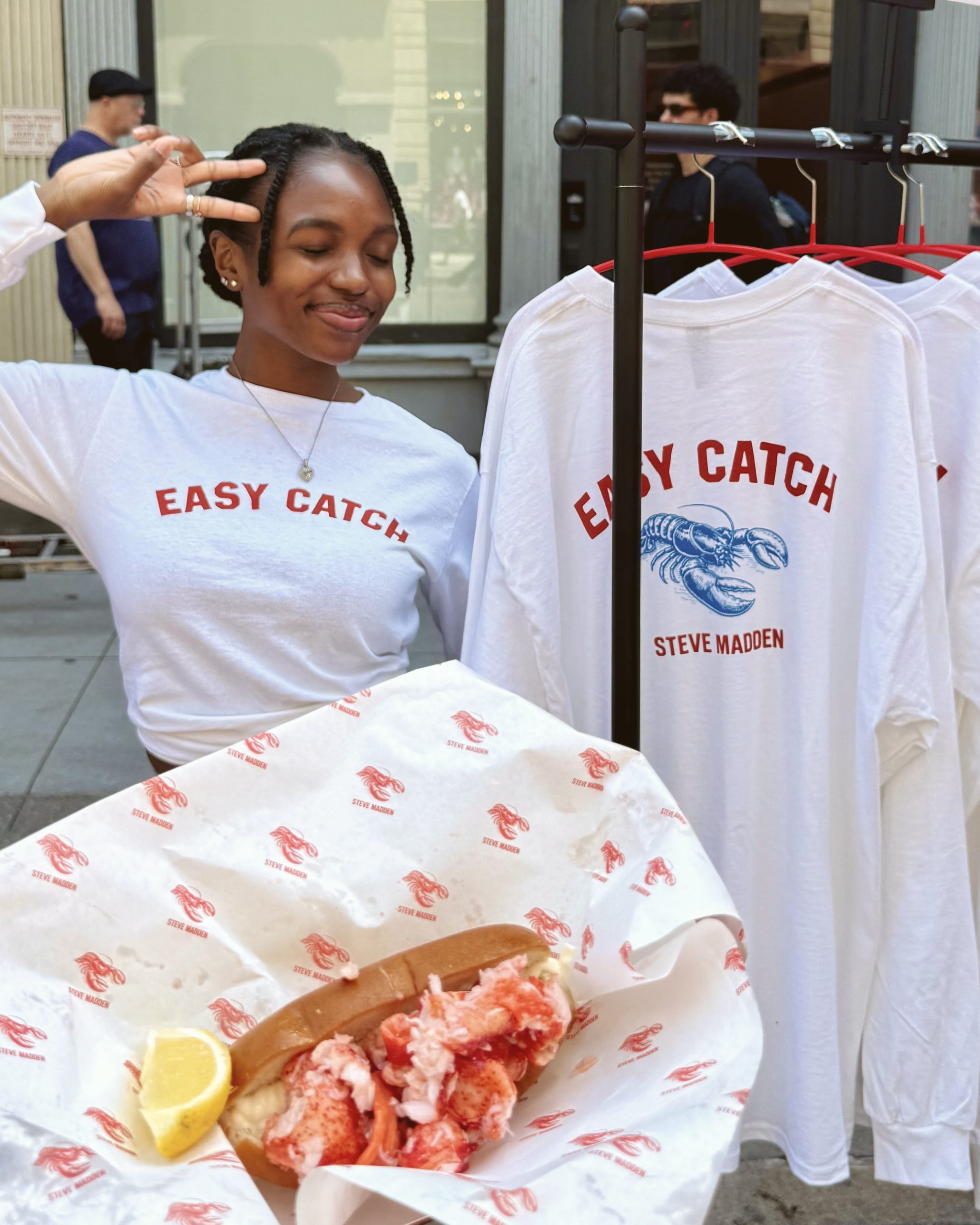 Person in EASY CATCH shirt flashes peace sign by a rack; lobster roll with lemon in front.